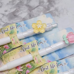 May include: White pens with flower-shaped toppers in yellow, pink, and white, resting on cards with the text "Best Life Ever." The cards feature a floral and sky design. The pens are a stationery item.