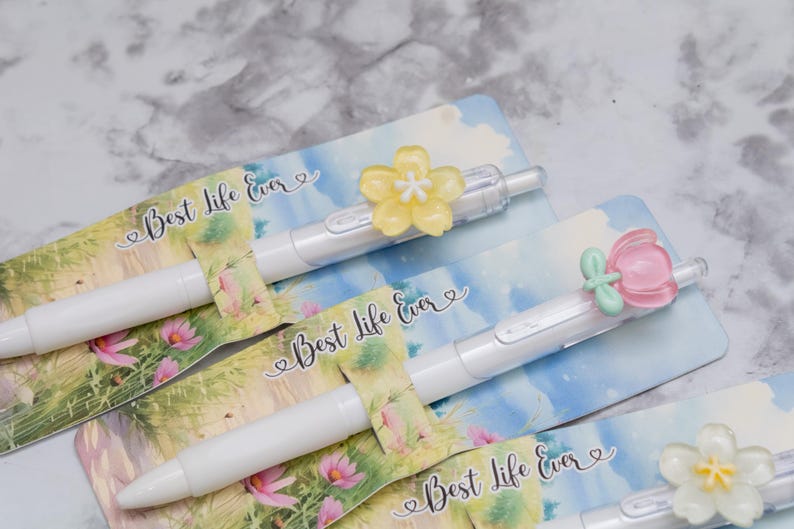 May include: White pens with flower-shaped toppers in yellow, pink, and white, resting on cards with the text "Best Life Ever." The cards feature a floral and sky design. The pens are a stationery item.