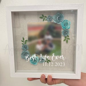 May include: White shadow box with a photo inside and a blue paper flower wreath around the photo. The text "Best Life Ever" and the date "11.12.2023" are written below the photo.