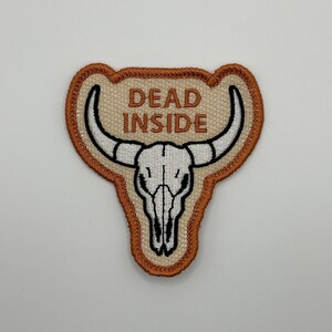 May include: Embroidered patch featuring a white bull skull with black details and curved horns. The patch has the words "DEAD INSIDE" in capital letters above the skull. The patch is bordered with a brown and tan design.