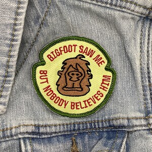 May include: A green, yellow, and red embroidered patch with the text "BIGFOOT SAW ME BUT NOBODY BELIEVES HIM". The patch features a cartoon image of Bigfoot with brown fur and a red nose.