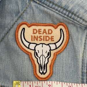 May include: Embroidered patch with a tan background, a white bull skull, and the words "DEAD INSIDE" in brown. The patch is outlined in brown and tan, and is approximately 2 inches wide.