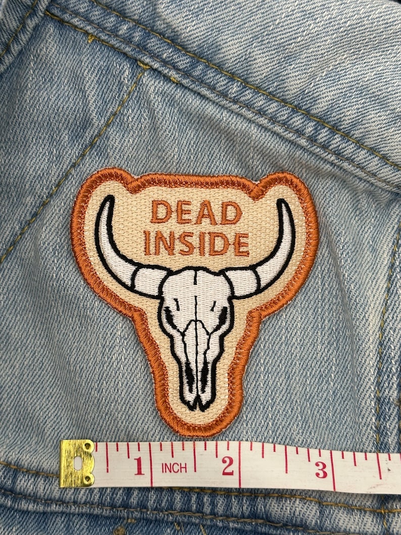 May include: Embroidered patch with a tan background, a white bull skull, and the words "DEAD INSIDE" in brown. The patch is outlined in brown and tan, and is approximately 2 inches wide.