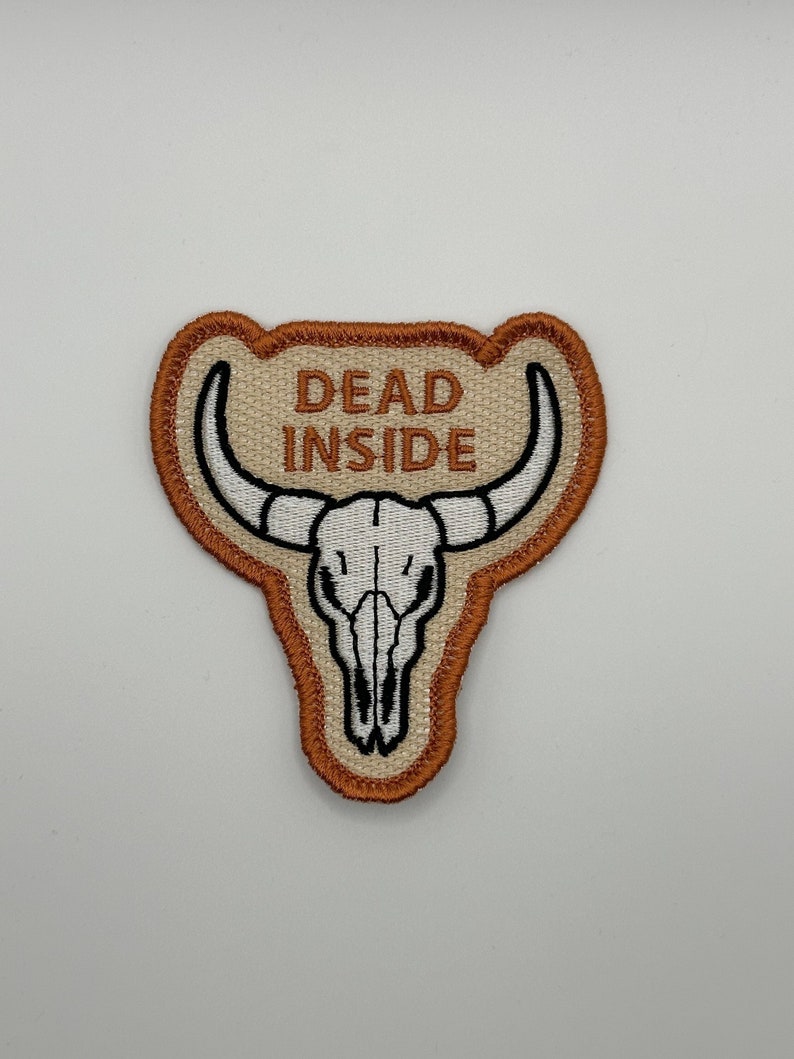 May include: Embroidered patch featuring a white bull skull with black details and curved horns. The patch has the words "DEAD INSIDE" in capital letters above the skull. The patch is bordered with a brown and tan design.