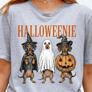 May include: A heather gray short-sleeve t-shirt featuring the orange text HALLOWEENIE above an illustration of three dachshund dogs. One dog wears a witch hat and cape, one is covered by a white ghost sheet, and one wears a witch hat and pumpkin.