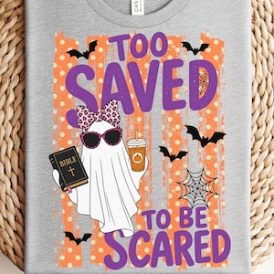 May include: A heather gray t-shirt featuring a graphic of a white ghost wearing pink leopard print bow and sunglasses, holding a black Bible and a coffee cup. The text reads TOO SAVED TO BE SCARED, surrounded by orange polka dots, bats, and a spider web.