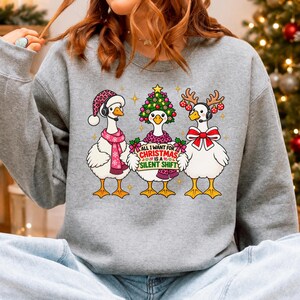 May include: A heather grey sweatshirt featuring a graphic of three white geese. The left goose wears a pink leopard print hat and scarf. The centre goose wears a Christmas tree hat and holds a sign reading All I Want For Christmas Is A Silent Shift. The right goose wears reindeer antlers and a red bow.
