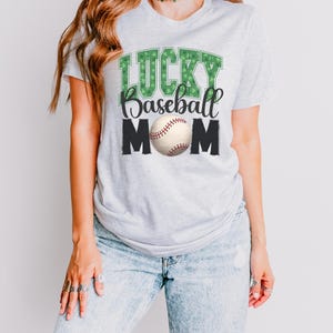 Puede incluir: Una camiseta gris con el texto "LUCKY Baseball MOM" en negro y verde. La palabra "LUCKY" está formada por tréboles verdes. Una pelota de béisbol blanca con costuras rojas está en el centro del texto.