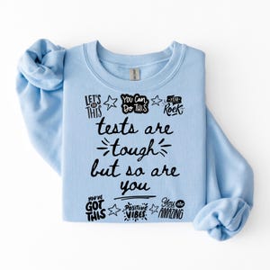May include: A pale blue sweatshirt featuring black text. The text includes motivational phrases such as "Tests are tough but so are you", "You've got this", and "You are amazing".