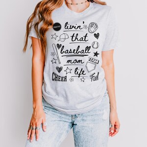 May include: A grey t-shirt with a baseball theme. The shirt has black text that says "Livin' that baseball mom life" and is surrounded by baseball-themed illustrations.
