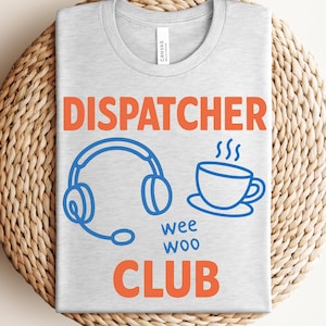 Puede incluir: Camiseta gris claro con la palabra "DISPATCHER" en naranja. Debajo, un gráfico azul de auriculares y una taza de café humeante. Las palabras "wee woo CLUB" también son de color naranja. La camiseta está doblada sobre una superficie tejida.