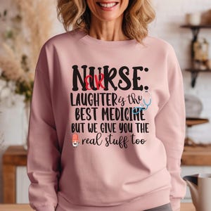 Może przedstawiać: Jasnoróżowa bluza z napisem "NURSE" w kolorze czarnym, litera "R" w kolorze czerwonym. Poniżej tekst "LAUGHTER is the BEST MEDICINE BUT WE GIVE YOU THE real stuff too" ze stetoskopem i pigułką.