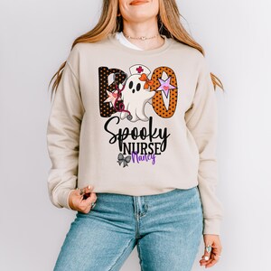 Może przedstawiać: Beżowa bluza z grafiką o tematyce Halloween. Projekt zawiera słowo "BOO" z pomarańczowymi i czarnymi literami w kropki, ducha w czepku pielęgniarki oraz słowa "Spooky Nurse Nancy".