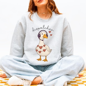Puede incluir: Sudadera gris claro con un dibujo de un ganso con auriculares y un collar de corazones. La palabra "dispatcher" está escrita encima del ganso en una fuente de escritura. El ganso tiene pico y patas naranjas.