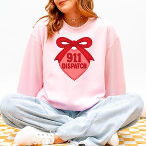 Puede incluir: Sudadera rosa claro con un lazo rojo y un gráfico en forma de corazón. El gráfico dice "911 DISPATCH" en rojo. La sudadera es de manga larga y tiene cuello redondo.