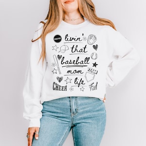 Op de afbeelding: Witte trui met zwarte tekst en illustraties die "Livin' that baseball mom life" zeggen. De illustraties omvatten honkballen, knuppels, harten, sterren en een honkbalhandschoen.