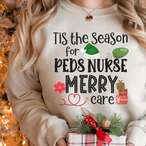 Puede incluir: Sudadera color crema con el texto "Tis the Season for Peds Nurse Merry Care". El diseño incluye un gorro de Papá Noel verde, un copo de nieve rojo y blanco, un estetoscopio y regalos envueltos. Un tema festivo navideño.