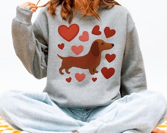 Op de afbeelding: Een grijze sweatshirt met een bruine teckel en rode en roze harten. De hond is omringd door harten van verschillende groottes. De sweatshirt heeft lange mouwen en een ronde hals.