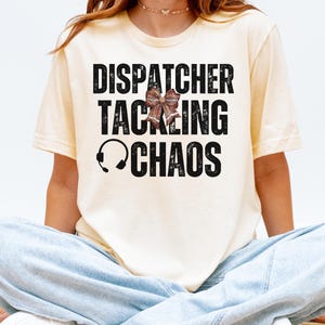 May include: A light yellow t-shirt with the words "DISPATCHER TACKLING CHAOS" in large, distressed black letters. A headset graphic and a decorative bow are also featured. The shirt is worn by a person.