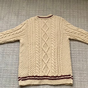May include: Cream-colored cable-knit sweater with a crew neck and long sleeves. The sweater features a diamond pattern down the front and cable knit detailing on the sleeves. The cuffs and hem have maroon stripes.