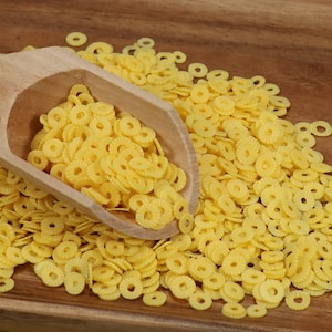 May include: A wooden scoop filled with yellow pasta shaped like small, flat, flower-like rings. The pasta is scattered on a wooden surface.