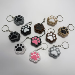 May include: A collection of ten paw print keychains in various colors, including black, white, gray, brown, pink, and beige. Each keychain features a 3D paw print design with a contrasting color for the paw pads.