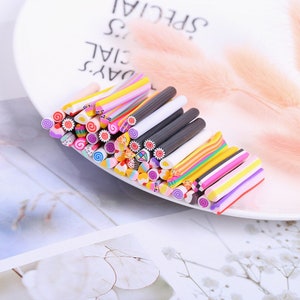 May include: Assortment of colorful polymer clay sculpting tools. The tools are in the shape of small sticks with various designs and patterns on the ends. The tools are arranged in a fan shape on a white plate.