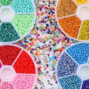 May include: A collection of colorful seed beads in a variety of colors, including turquoise, green, red, pink, gold, and blue. The beads are organized in a multi-compartment container and scattered on a white surface.