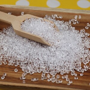 May include: A wooden scoop filled with clear, square-shaped plastic beads on a wooden surface.