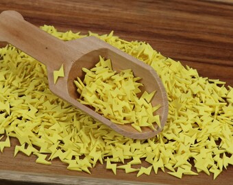 May include: A wooden scoop filled with yellow lightning bolt shaped sprinkles. The sprinkles are scattered on a wooden surface.