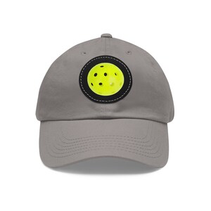 May include: A gray baseball cap with a black and white circular patch featuring a yellow pickleball.