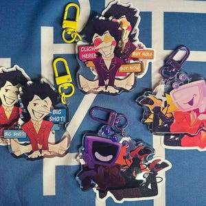 以下が含まれることがあります： スーツを着た漫画のキャラクターが描かれた、カラフルなアクリルキーホルダーのコレクションです。「BIG SHOT!」と「CLICK HERE! BUY NOW」という文字が入ったキーホルダーもあります。キーホルダーには黄色と紫の留め金が付いています。