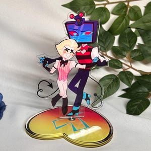 Może przedstawiać: Przezroczysta figurka akrylowa przedstawiająca dwie tańczące postacie z kreskówek. Jedna postać ma głowę w kształcie ekranu telewizora, druga jest blondynką i ma na sobie różowy strój. Podstawa w kształcie serca ma gradient żółto-pomarańczowo-czerwony.