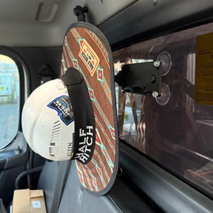 Könnte beinhalten: Ein braun-beiger Half Hitch-Hut mit geometrischem Muster und dem Text "HALF HITCH" ist an einer schwarzen Halterung mit Saugnäpfen befestigt. Ein weißer Schutzhelm mit Aufklebern hängt darunter.