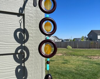 May include: A wooden wind chime with three circular rings, each containing a yellow-orange oval-shaped bead. The rings are connected by turquoise beads and a brown wooden bead. A bronze bell hangs from the bottom.