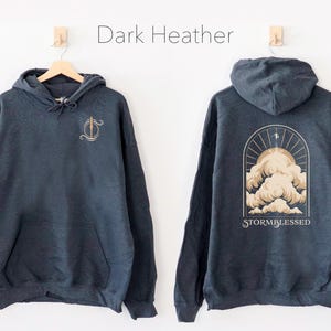 May include: A dark heather grey hoodie with a graphic of a stylized cloud with the text "STORMBLESSED" on the back. The front of the hoodie has a small graphic of a stylized sword.