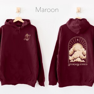 May include: A maroon hoodie with a graphic of a stylized cloud with the text "STORMBLESSED" on the back. The front of the hoodie has a small graphic of a sword.