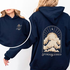 May include: A navy blue hoodie with a graphic of a sun and clouds on the back. The text "STORMBLESSED" is printed below the graphic.