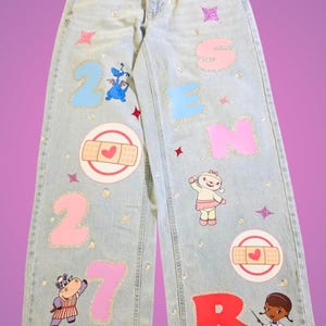 May include: Light-wash denim jeans featuring glittery numbers and letters spelling 2027 SENIOR. The pants are decorated with cartoon characters, adhesive bandage graphics with hearts, and small sparkling star accents scattered across the fabric.