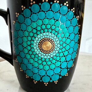 May include: A black ceramic mug with a teal and gold mandala design. The mandala is painted in a circular pattern with dots of varying sizes.
