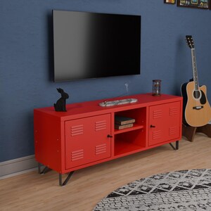以下が含まれることがあります： 2つのキャビネットドアと中央のオープンシェルフを備えた赤いテレビ台。スタンドは、通気孔のディテールを備えた金属製のロッカー風のデザインです。黒いテレビがスタンドの上に設置されています。黒いウサギの置物が上にあります。