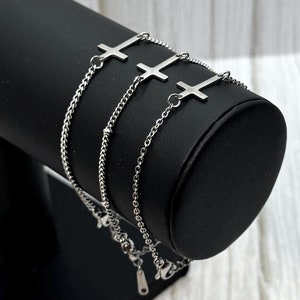 May include: Three silver chain bracelets with cross charms. The bracelets are displayed on a black jewellery stand.