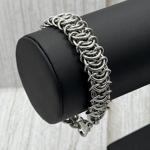 Könnte beinhalten: Ein silbernes Kettenhemdarmband mit einem Verschluss. Das Armband wird auf einem schwarzen Ständer präsentiert.