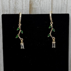 May include: Gold earrings with a delicate branch design featuring green leaves and a clear crystal dangle.