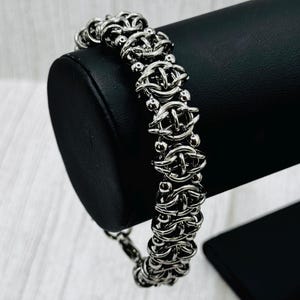 May include: A silver-colored chainmail bracelet with a detailed, interwoven design. The bracelet is displayed on a black stand, showcasing its intricate links and polished finish. The jewelry piece is a stylish accessory.