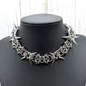 May include: A silver choker necklace with a spiked design. The necklace features a chainmail-style construction with star-shaped accents and sharp spikes. The necklace is displayed on a black mannequin.
