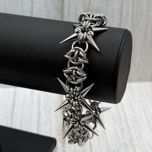 May include: A silver bracelet with a chain-link design, featuring multiple star-shaped spikes. The bracelet is displayed on a black stand against a light-coloured wooden background. The spikes are evenly spaced along the bracelet.