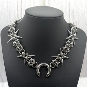 Op de afbeelding: Een zilverkleurige ketting met een stekelig ontwerp. De ketting heeft meerdere stervormige elementen met scherpe spikes en een centrale hoefijzervormige hanger met naar beneden gerichte spikes. De ketting wordt getoond op een zwarte mannequin.