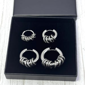 May include: A set of four silver-toned hoop earrings in a black box. The earrings are of varying sizes, each featuring multiple small, circular accents. The box is open, revealing the earrings against a dark interior.