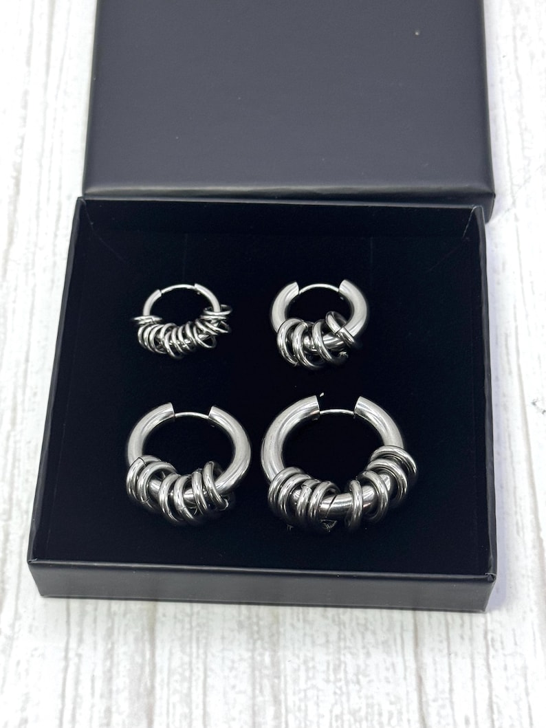 May include: A set of four silver-toned hoop earrings in a black box. The earrings are of varying sizes, each featuring multiple small, circular accents. The box is open, revealing the earrings against a dark interior.